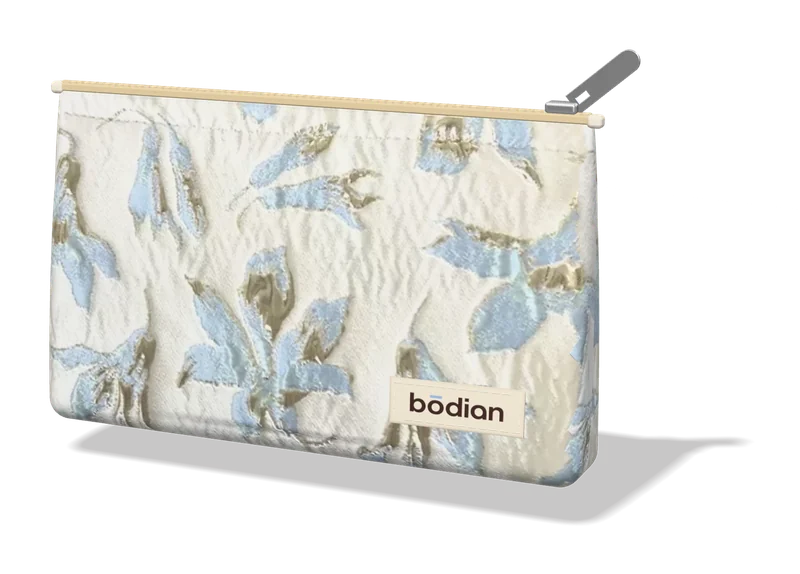 The free limited-edition pouch: ivory jacquard woven with pale blue and gold lilies, a beige zip with a silver pull, and the bōdian label at its bottom right